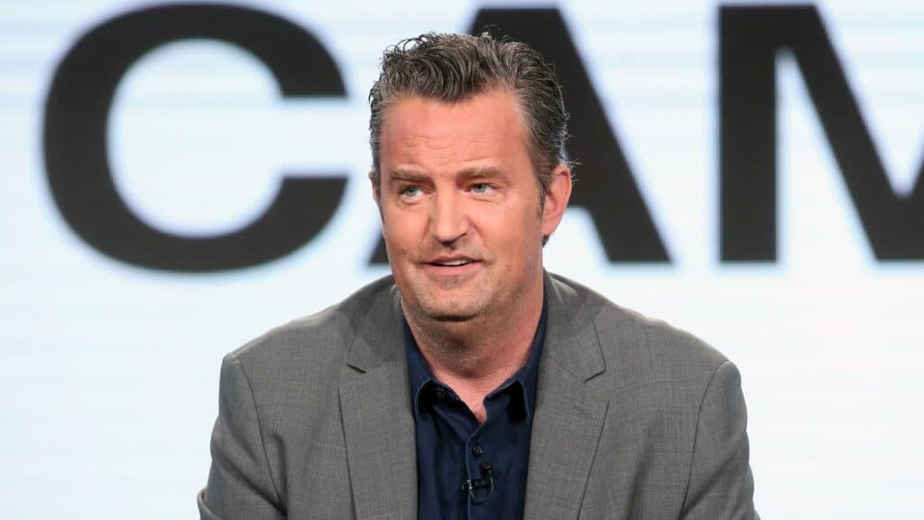 Matthew Perry durante una presentación en un evento, vestido con traje gris y camisa negra, reflejando su carrera en la actuación.