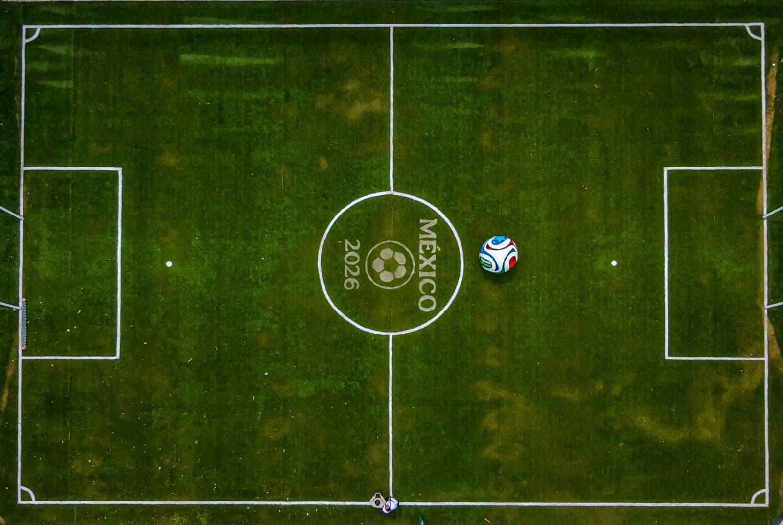Campo de fútbol con el logo de México 2026, destacando un balón en el centro y líneas de juego bien definidas.