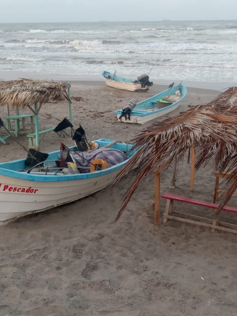 Botes de pesca en la playa de Playa Hermosa, con sombrillas de palma y olas del mar al fondo, reflejando el entorno turístico afectado.