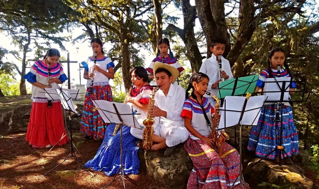 Niños y jóvenes en trajes tradicionales tocan instrumentos musicales en un entorno natural, promoviendo la educación musical en comunidades.