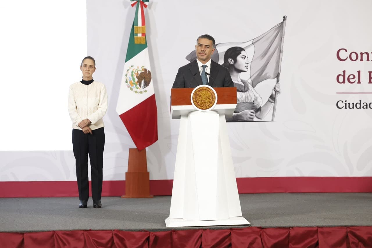 El funcionario ofrece un discurso en un evento oficial, con la bandera de México y un retrato histórico de fondo.