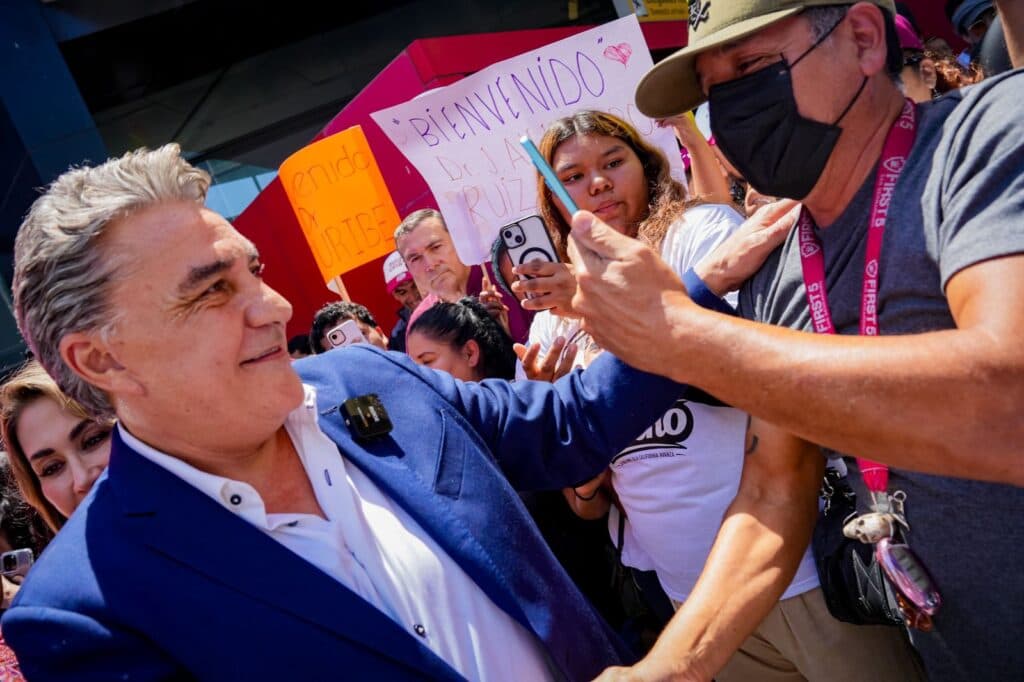 Hombres y mujeres de izquierda en Baja California apoyan a Jesús Alejandro Ruiz Uribe durante un evento con pancartas de respaldo.