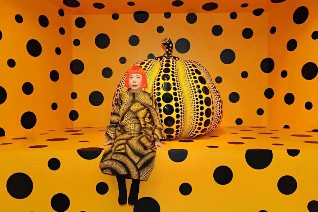 Yayoi Kusama sentada en un entorno amarillo con lunares negros, junto a una calabaza decorada con el mismo patrón,