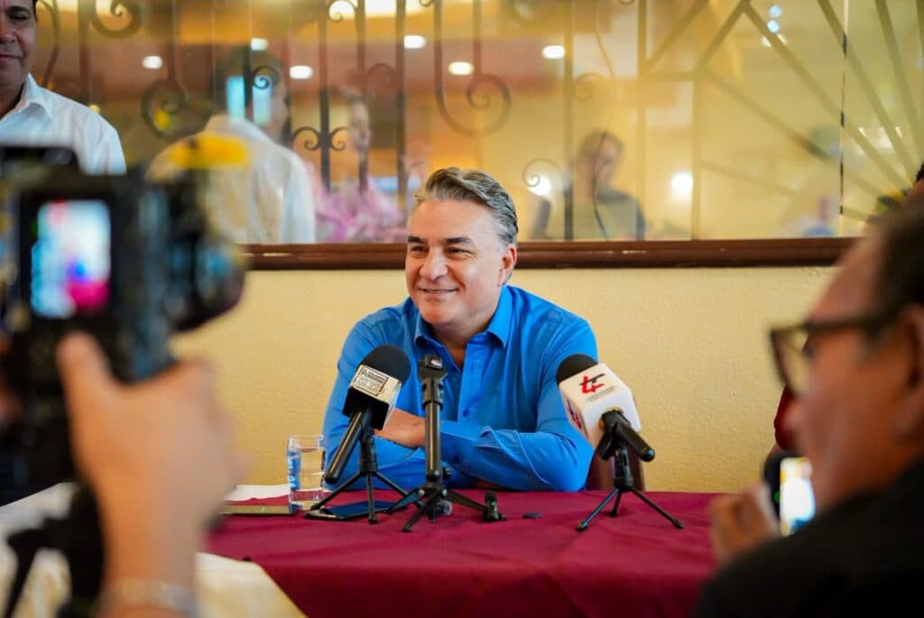 Jesús Ruiz Uribe sonríe durante una conferencia de prensa sobre las asambleas de MORENA en Baja California, rodeado de micrófonos y periodistas.