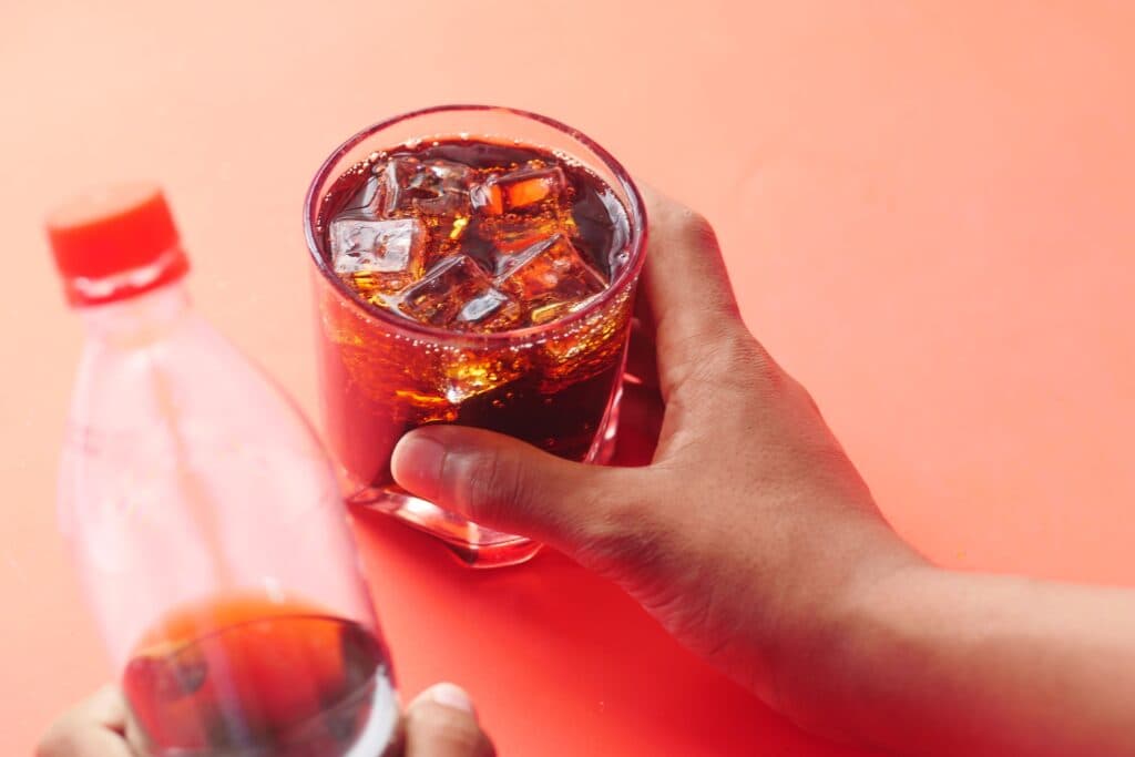 Vaso con refresco y hielo en una mano, mientras se sostiene una botella de bebida azucarada, resaltando el riesgo para la salud del hígado.