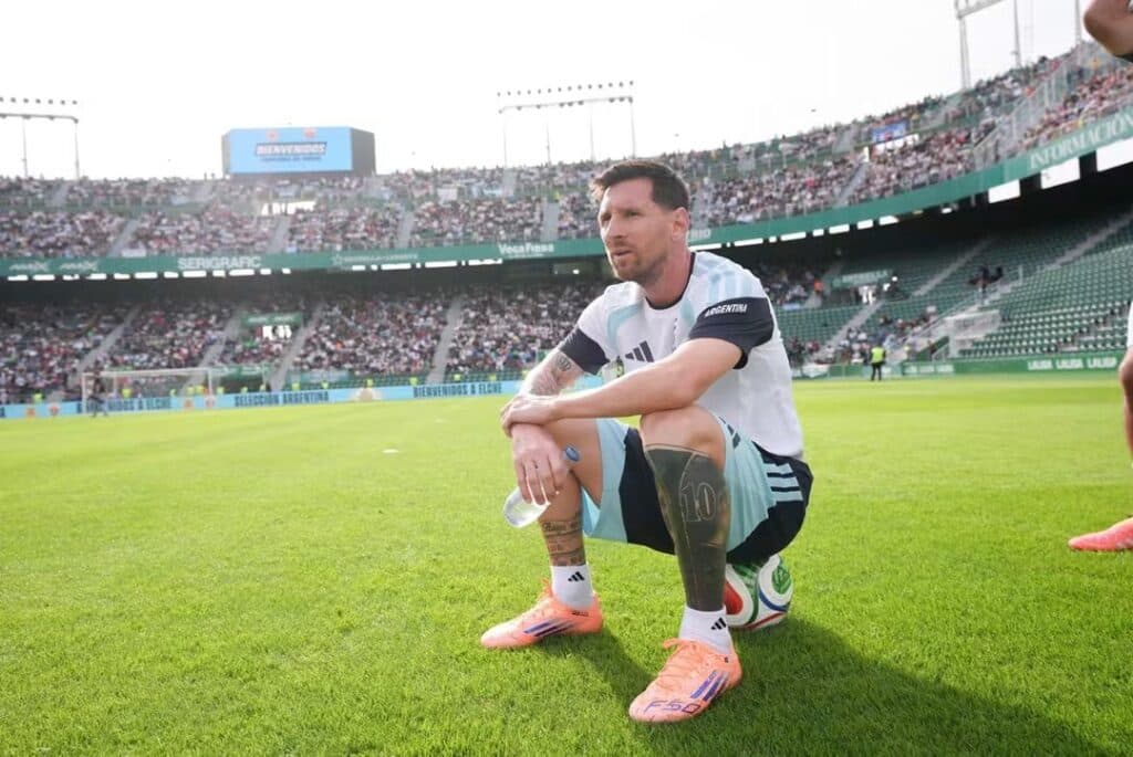 Lionel Messi sentado en el campo de juego, con la camiseta de la Selección Argentina, durante un partido lleno de aficionados.
