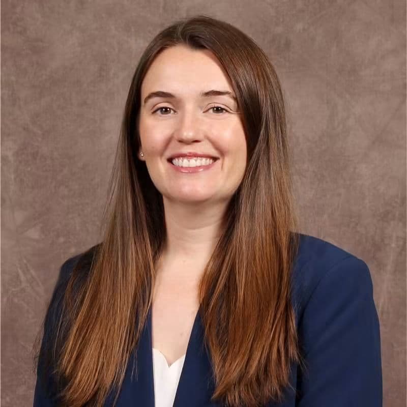 Mujer sonriente con cabello largo y castaño, vestida con un blazer oscuro, en un fondo neutro, identificada como