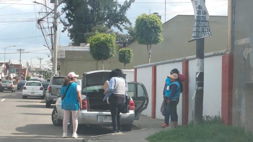 Encuestadores de Morena en Tlaxcala preparan cuestionarios en la calle, mientras vehículos pasan por el fondo.