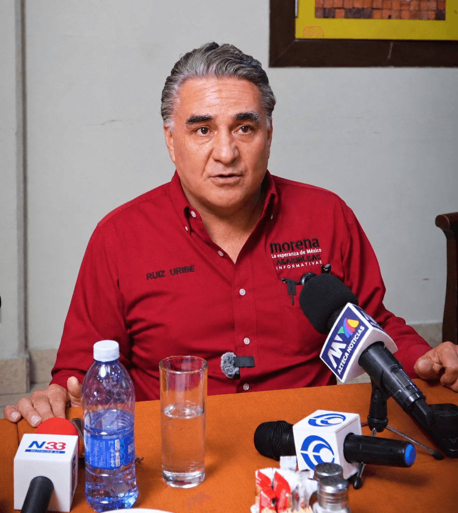 Ruiz Uribe habla ante medios sobre los logros de Claudia Sheinbaum, con micrófonos y agua en la mesa.