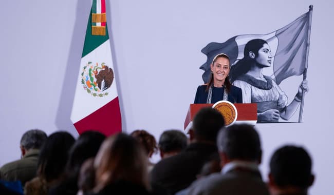 Claudia Sheinbaum en conferencia de prensa matutina en Palacio Nacional, con bandera de México y mural de mujer indígena de fondo.