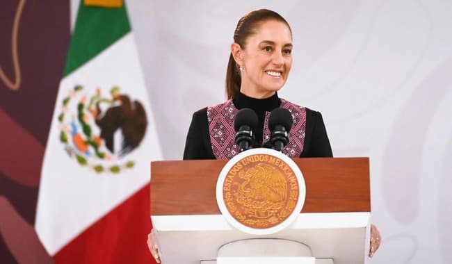 Claudia Sheinbaum sonríe durante su conferencia en Irapuato, Guanajuato, con la bandera de México de fondo, hablando sobre acuerdos comerciales.
