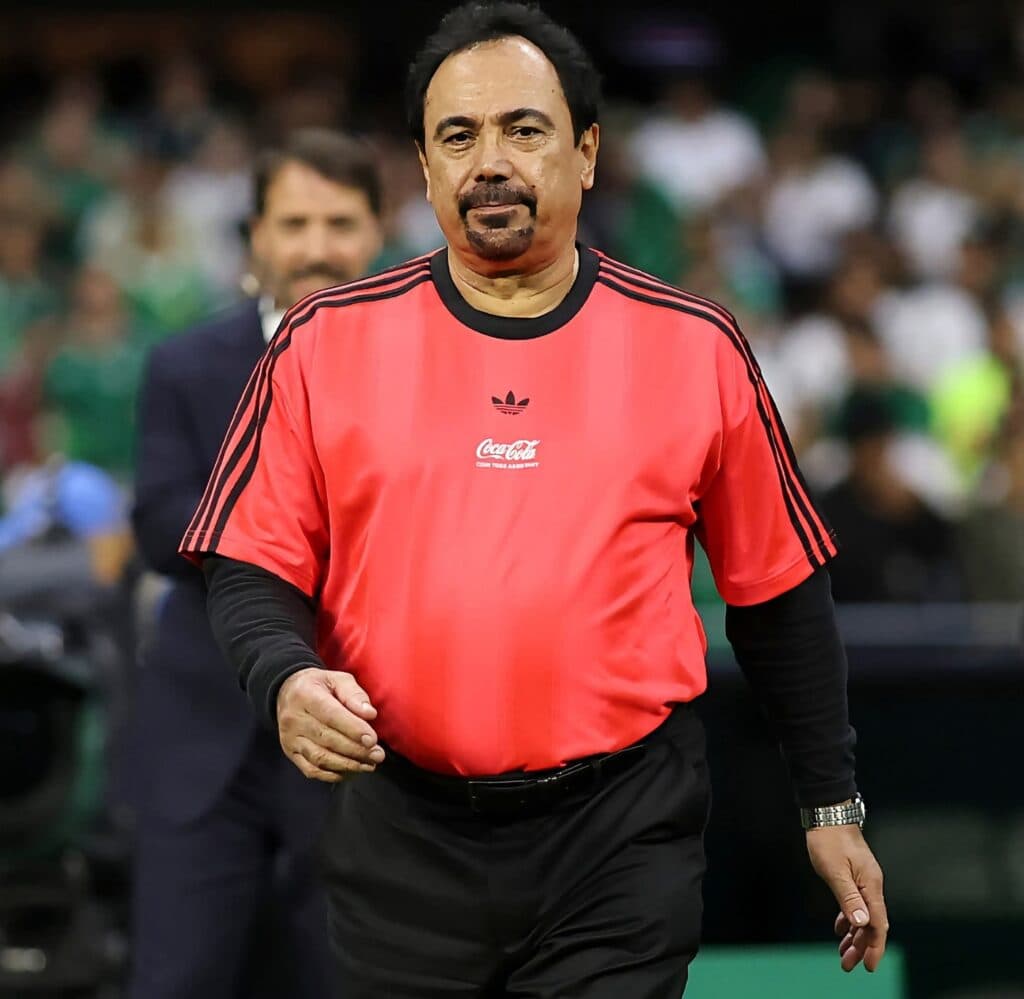 Hugo Sánchez vestido con camiseta roja de árbitro, caminando en un evento deportivo, discutiendo sobre el VAR en la Liga MX.