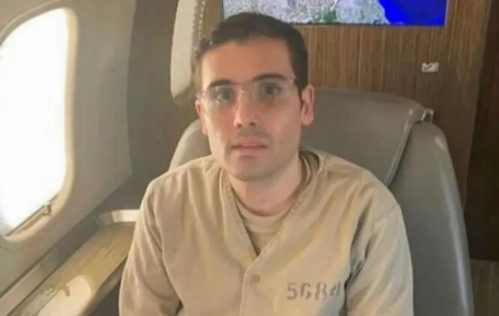Ovidio Guzmán López, hijo de Joaquín "El Chapo", sentado en un avión, vestido con un uniforme carcelario, en un contexto judicial.