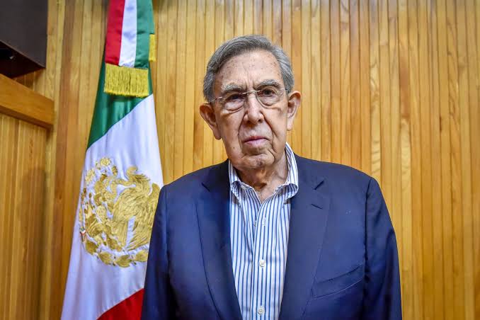 Cuauhtémoc Cárdenas, político y fundador del PRD, posando frente a la bandera de México en un evento oficial.